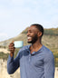 A dark-skinned man in a heather blue shirt, smiling and holding a light blue mug of tea, with bluffs in background.