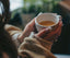 A dark-haired, light-skinned woman in a sweater holding a cup of tea in both hands.