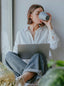 A light-skinned woman in a white button down and jeans, holding a laptop in her lap and drinking from a blue mug of tea.